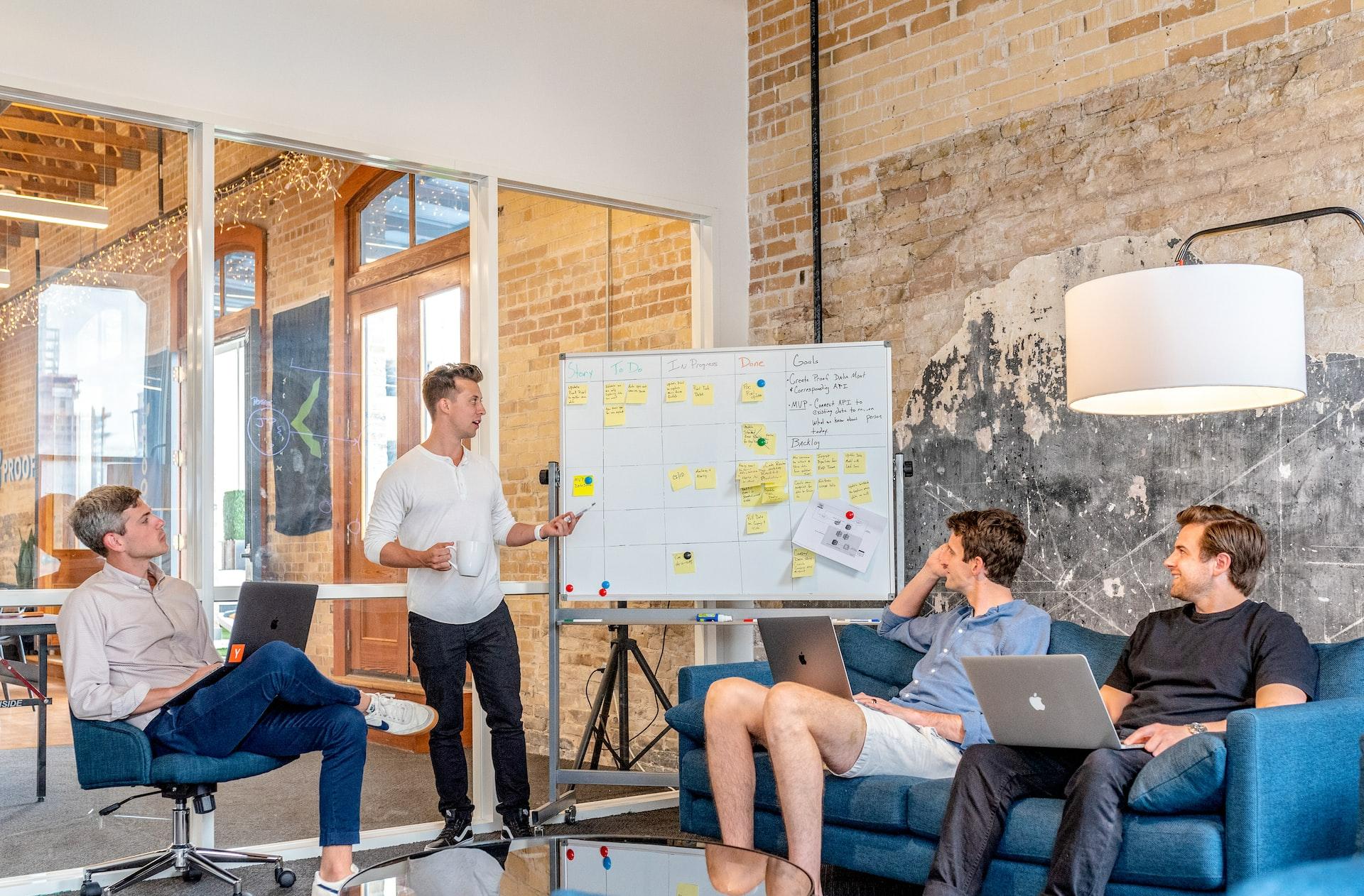 three men sitting while using laptops and watching man beside whiteboard